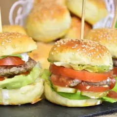 Mini burgery z sezamem, sałatą, pomidorem i sosem na czarnej desce.