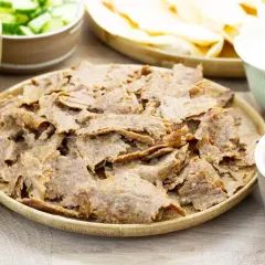 Pyszny, domowy kebab z piekarnika na talerzu, otoczony świeżymi dodatkami: poszatkowaną kapustą, pomidorami z ogórkiem, sosem i plackami tortilli.