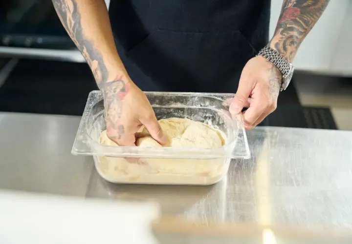Ręce z tatuażami mieszają ciasto na pizzę w pojemniku. Dowiedz się, jak szybko rozmrozić ciasto na pizzę.