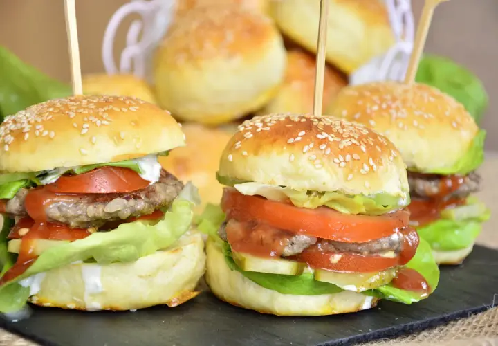 Mini burgery z sezamem, sałatą, pomidorem i sosem na czarnej desce.
