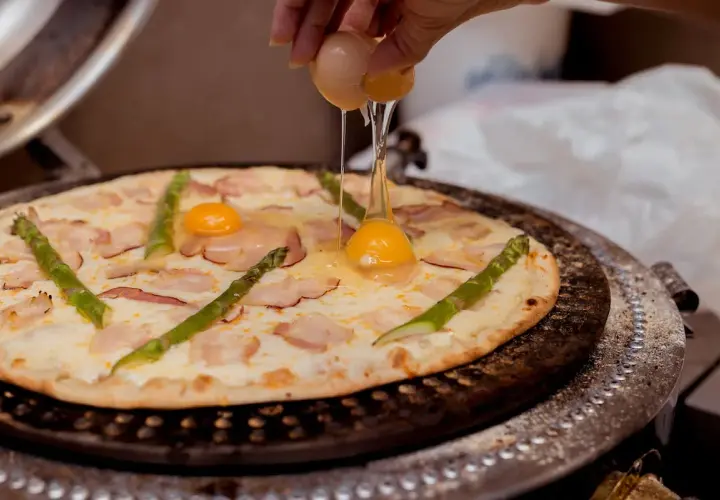 Ktoś dodaje jajko do pizzy z boczkiem i szparagami. Pyszna pizza z jajkiem w przygotowaniu.