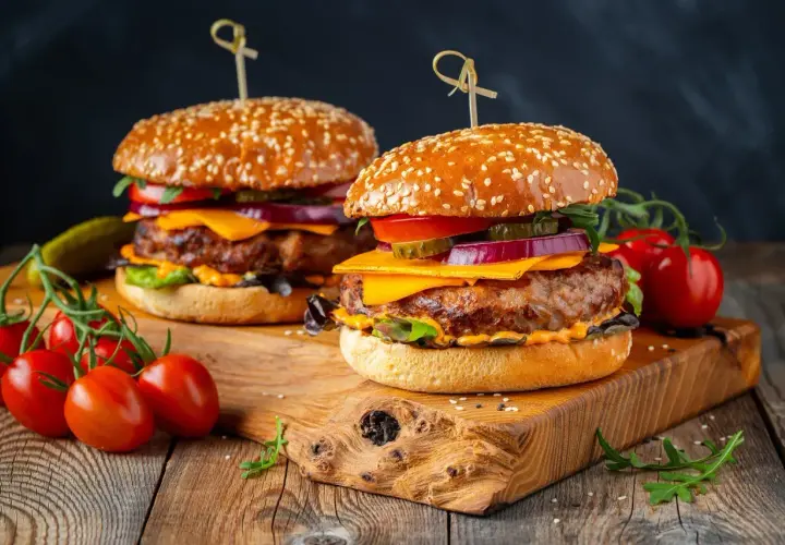 Dwa soczyste burgery z serem, warzywami i sezamową bułką na drewnianej desce.