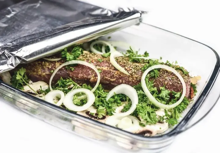 Mięso w naczyniu żaroodpornym, otoczone cebulą i ziołami, przykryte folią aluminiową która strona na grilla.