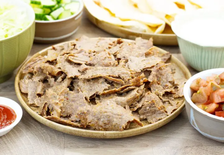 Pyszny, domowy kebab z piekarnika na talerzu, otoczony świeżymi dodatkami: poszatkowaną kapustą, pomidorami z ogórkiem, sosem i plackami tortilli.