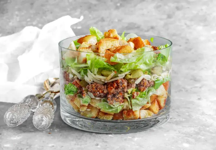 Warstwowa sałatka a'la Big Mac w szklanej misie, z mielonym mięsem, sałatą, serem, ogórkiem i grzankami.