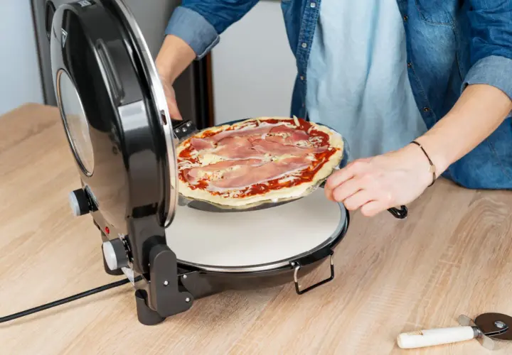 Osoba wkłada do piecyka okrągłą pizzę z szynką i serem, gotową do pieczenia. To będzie pyszna pizza Dziuzeppe!