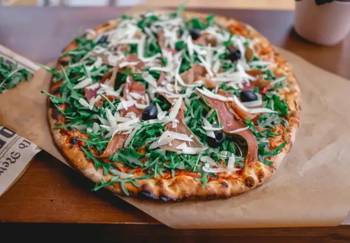 Apetyczna pizza prosciutto z rukolą, oliwkami i parmezanem, podana na papierze.