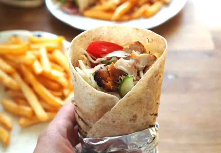 Ręka trzymająca prawdziwy kebab zawinięty w tortillę, z widocznym kurczakiem, warzywami i frytkami w tle.