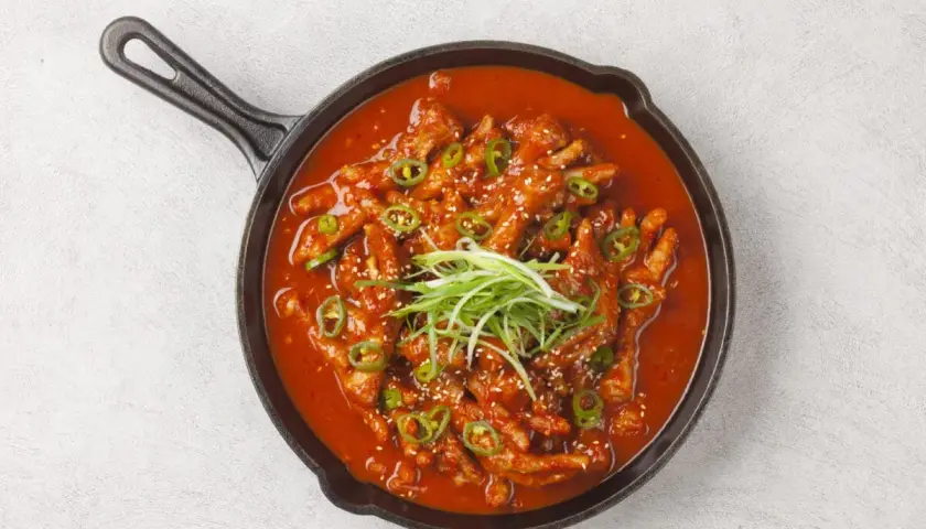 Kurczak w sosie gochujang, ozdobiony zieloną cebulką i sezamem, podany w żeliwnej patelni.