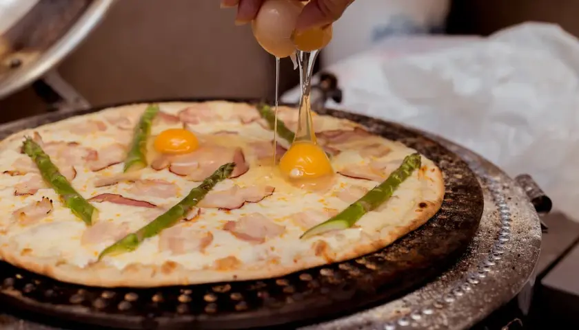 Ktoś dodaje jajko do pizzy z boczkiem i szparagami. Pyszna pizza z jajkiem w przygotowaniu.