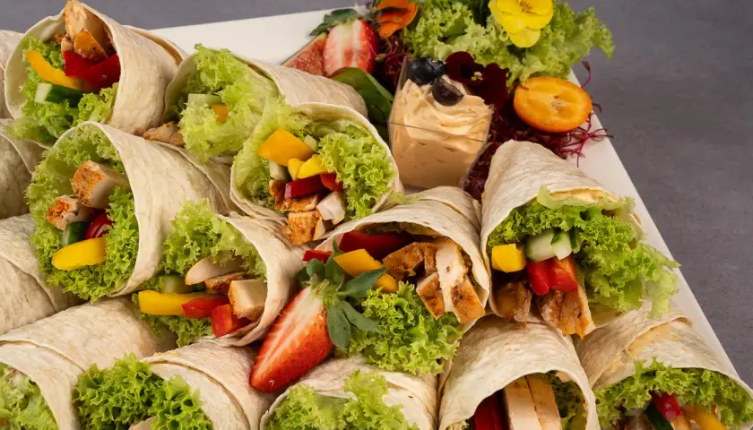 Mcwrap grillowany kurczak z warzywami i sałatą, ułożone na talerzu z dodatkami: dipem, owocami i ziołami.