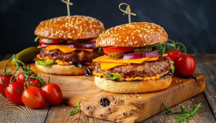 Dwa soczyste burgery z serem, warzywami i sezamową bułką na drewnianej desce.