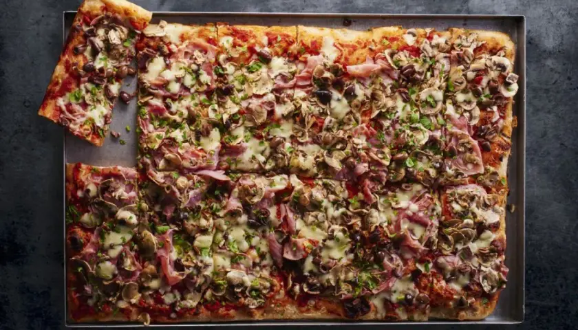 Najlepsza pizza domowa na dużą blachę, z szynką, pieczarkami i oliwkami, posypana świeżą natką pietruszki.