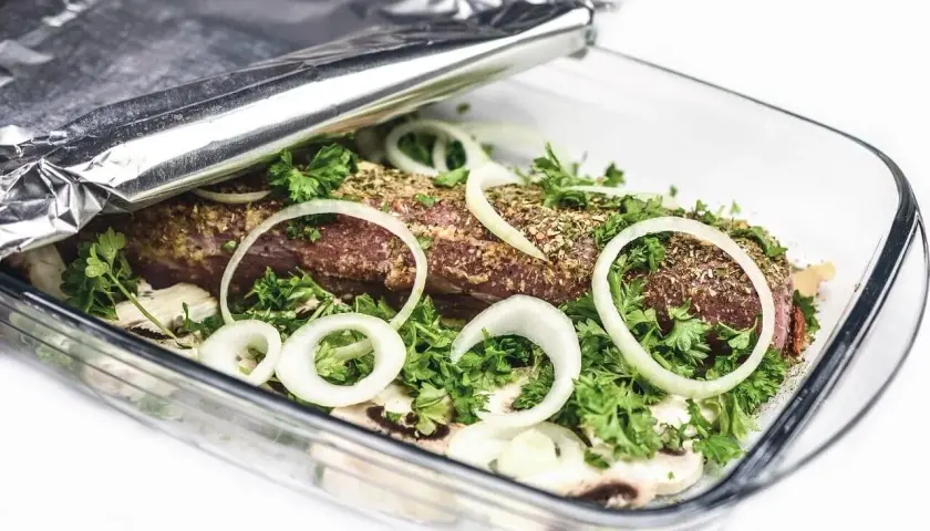 Mięso w naczyniu żaroodpornym, otoczone cebulą i ziołami, przykryte folią aluminiową która strona na grilla.