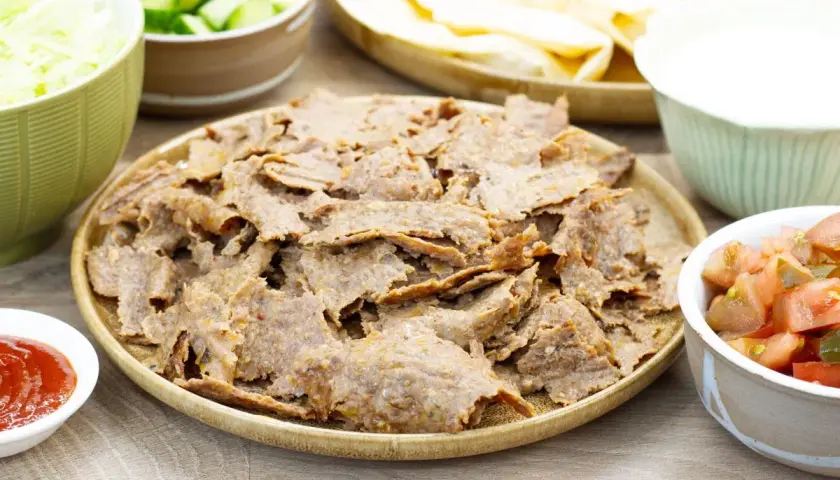 Pyszny, domowy kebab z piekarnika na talerzu, otoczony świeżymi dodatkami: poszatkowaną kapustą, pomidorami z ogórkiem, sosem i plackami tortilli.