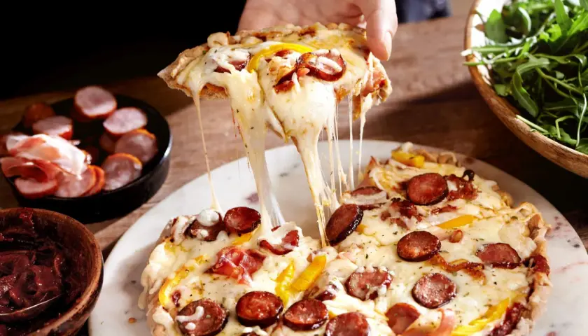 Rozciągający się ser z kawałka farmerskiej pizzy z kiełbasą i papryką. Obok miseczka z rukolą i wędlinami.