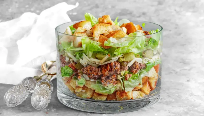 Warstwowa sałatka a'la Big Mac w szklanej misie, z mielonym mięsem, sałatą, serem, ogórkiem i grzankami.