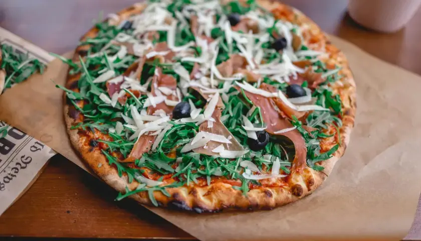 Apetyczna pizza prosciutto z rukolą, oliwkami i parmezanem, podana na papierze.