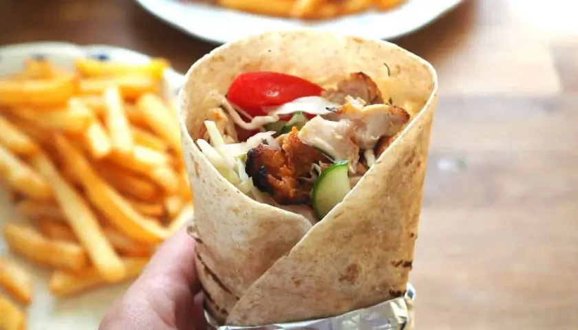 Ręka trzymająca prawdziwy kebab zawinięty w tortillę, z widocznym kurczakiem, warzywami i frytkami w tle.