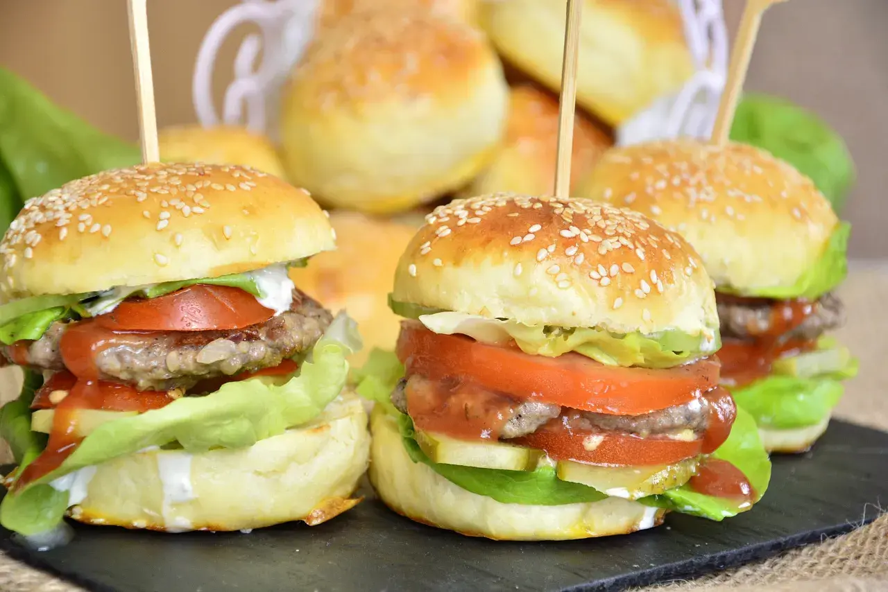 Mini burgery z sezamem, sałatą, pomidorem i sosem na czarnej desce.
