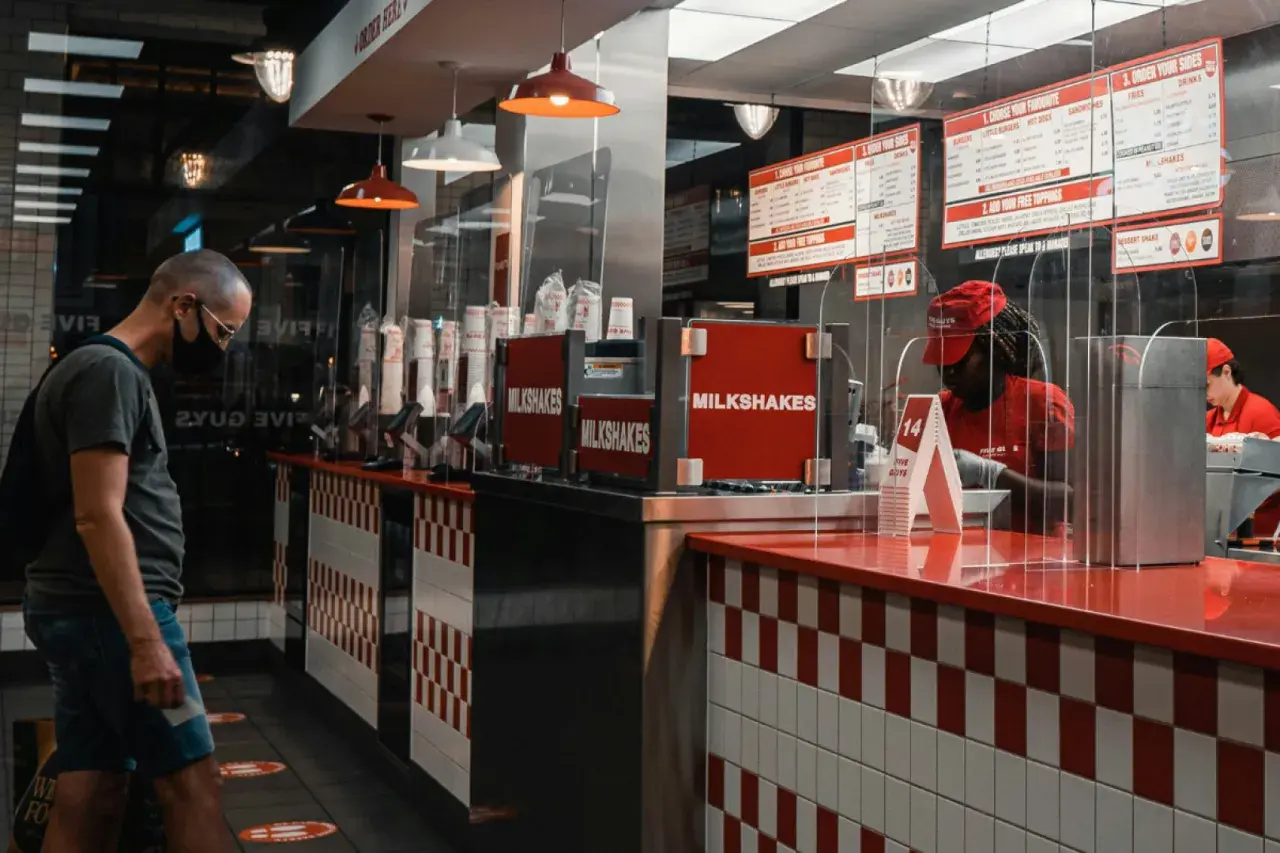 Klient w masce w Five Guys Polska czeka na zamówienie. W tle widać menu i pracownika za ladą.
