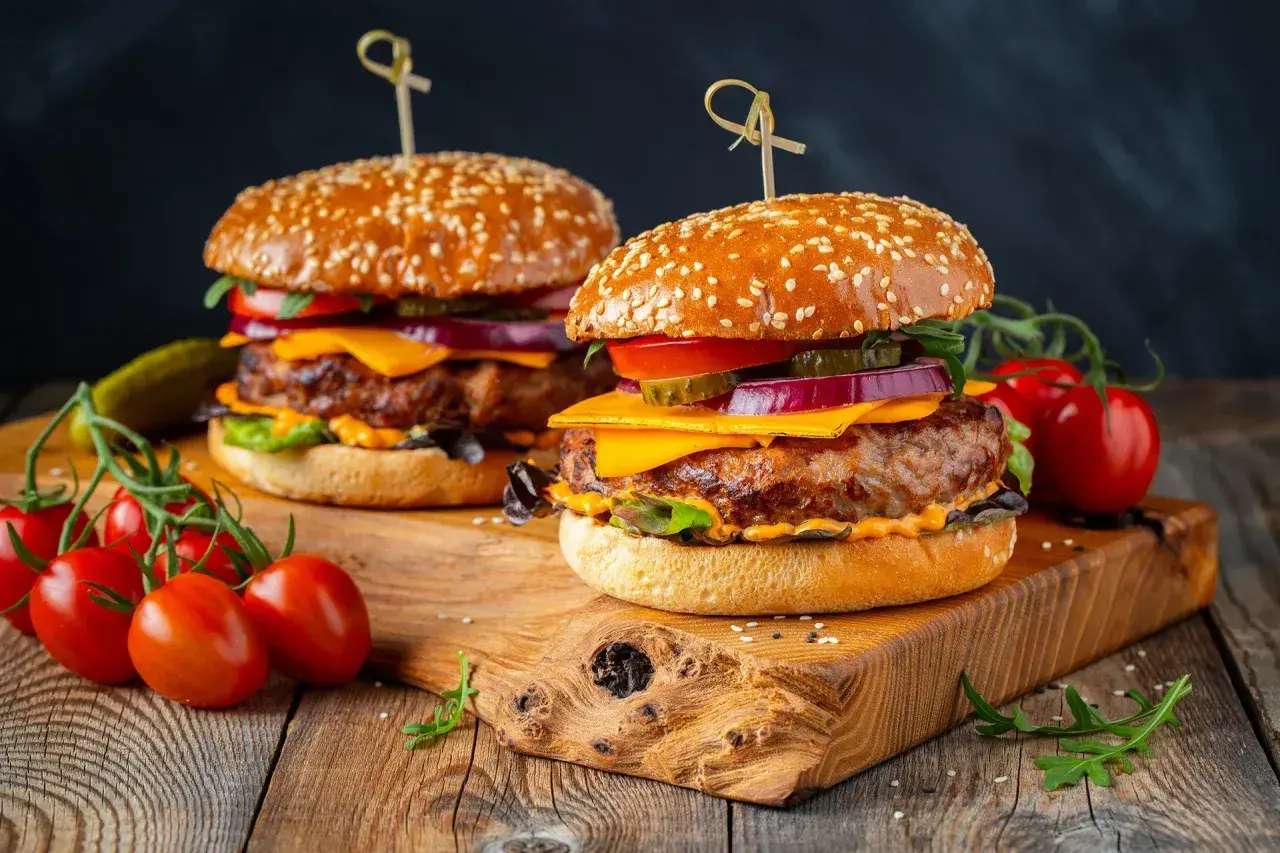 Dwa soczyste burgery z serem, warzywami i sezamową bułką na drewnianej desce.