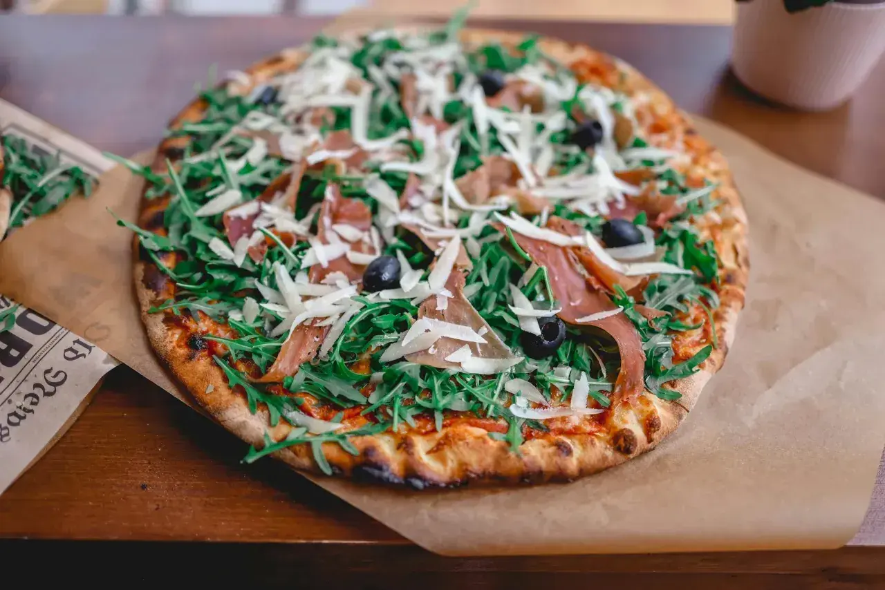 Apetyczna pizza prosciutto z rukolą, oliwkami i parmezanem, podana na papierze.